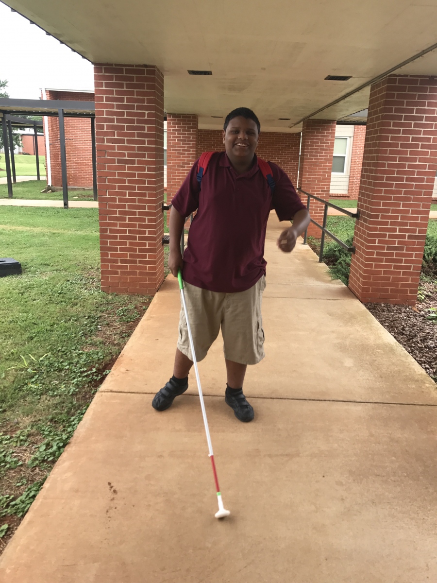 A male student walking on a sidewalk with a white cane. 